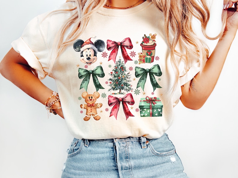 May include: A white t-shirt with a Christmas-themed design featuring Mickey Mouse, a gingerbread man, a Christmas tree, presents, and bows. The design is in a watercolor style with red, green, and brown colors.