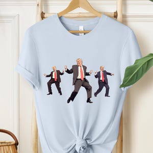 May include: Pale blue t-shirt with a graphic of three dancing figures in suits and red ties. The figures are in different poses, with one in the centre and two on either side. The t-shirt is hanging on a wooden hanger.