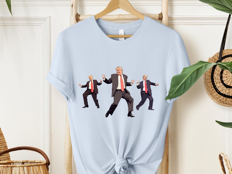 May include: Pale blue t-shirt with a graphic of three dancing figures in suits and red ties. The figures are in different poses, with one in the centre and two on either side. The t-shirt is hanging on a wooden hanger.
