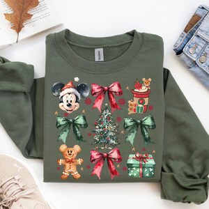 May include: A green sweatshirt with a Christmas-themed design featuring Mickey Mouse, a gingerbread man, a Christmas tree, and a cup of hot cocoa. The design is in a watercolor style with red, green, and gold accents.