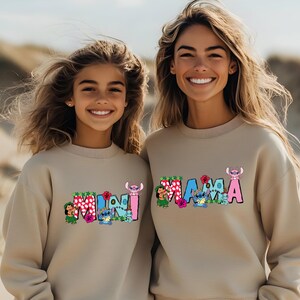 May include: Two people wearing beige sweatshirts with colorful graphic designs. The adult sweatshirt says "MAMA" and the child's sweatshirt says "MINI".