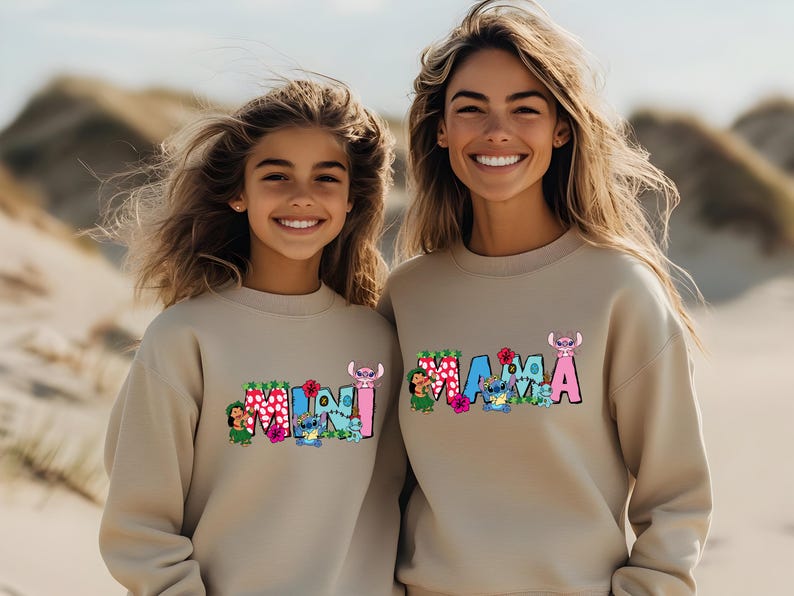 May include: Two people wearing beige sweatshirts with colorful graphic designs. The adult sweatshirt says "MAMA" and the child's sweatshirt says "MINI".