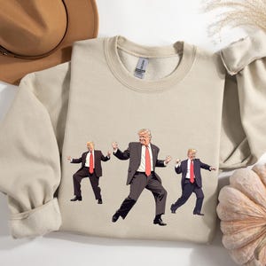May include: Beige crewneck sweatshirt with a graphic of three cartoon figures in suits and red ties, dancing. The sweatshirt is made of a soft material. A brown hat and a pumpkin are in the background.
