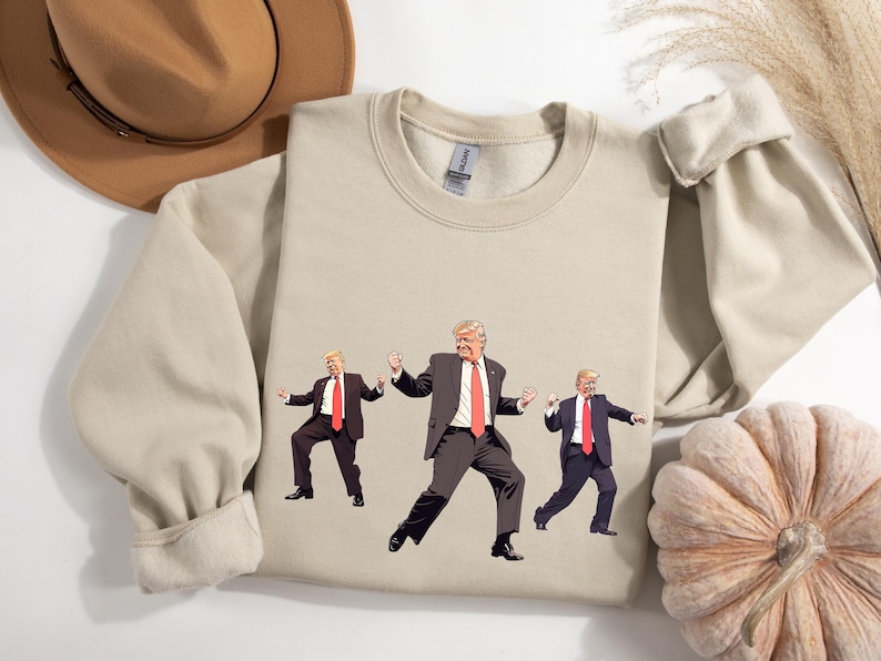 May include: Beige crewneck sweatshirt with a graphic of three cartoon figures in suits and red ties, dancing. The sweatshirt is made of a soft material. A brown hat and a pumpkin are in the background.