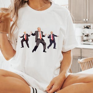 May include: White t-shirt with a graphic of three figures in suits and red ties in a dancing pose. The figures are in black suits with white shirts and red ties. The t-shirt is a casual style.
