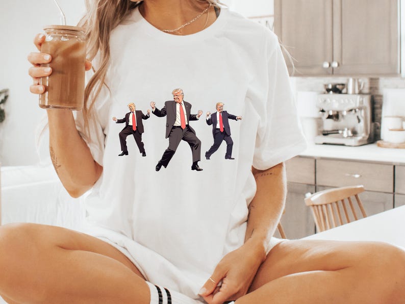 May include: White t-shirt with a graphic of three figures in suits and red ties in a dancing pose. The figures are in black suits with white shirts and red ties. The t-shirt is a casual style.