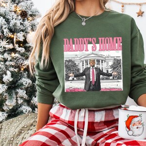 May include: A green sweatshirt with the text "DADDY'S HOME" above an image of a man in a suit with the White House in the background. The person is wearing red and white plaid pajama pants and holding a mug with a Santa Claus design.