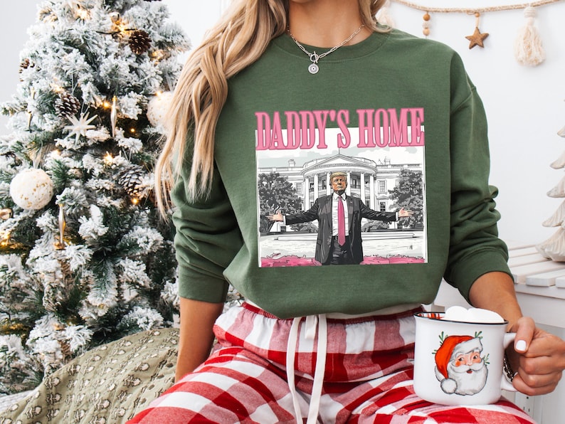 May include: A green sweatshirt with the text "DADDY'S HOME" above an image of a man in a suit with the White House in the background. The person is wearing red and white plaid pajama pants and holding a mug with a Santa Claus design.