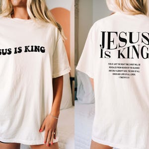 Puede incluir: Camiseta blanca de gran tama&ntilde;o con el texto "JESUS IS KING" impreso en negro en la parte delantera y un vers&iacute;culo b&iacute;blico en la parte posterior.