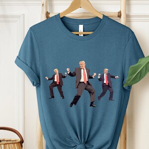 May include: A teal t-shirt with a graphic of three figures in suits and red ties, in a dancing pose. The figures are in a repeating pattern across the front of the shirt. The shirt is hanging on a wooden hanger.