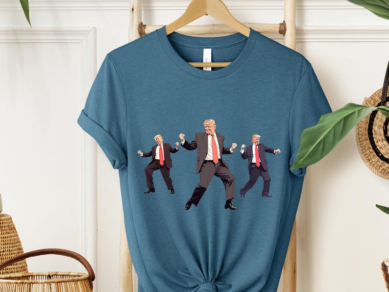 May include: A teal t-shirt with a graphic of three figures in suits and red ties, in a dancing pose. The figures are in a repeating pattern across the front of the shirt. The shirt is hanging on a wooden hanger.