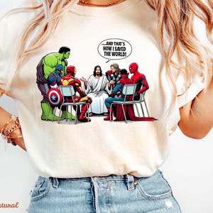 May include: Cream-colored t-shirt featuring a cartoon illustration of Jesus and superheroes. The image includes the Hulk, Captain America, Iron Man, Thor, Spider-Man, and Doctor Strange. Text in a speech bubble reads, "...AND THAT'S HOW I SAVED THE WORLD!"