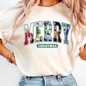 May include: A white t-shirt with a green and white "Merry Christmas" graphic featuring characters from popular Christmas movies. The graphic is in a collegiate style font.