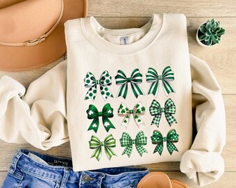 May include: A cream-colored sweatshirt with a green shamrock bow design. The bows are in various patterns, including polka dots, stripes, and plaid.