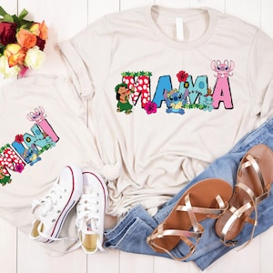 May include: Two white t-shirts with a cartoon character design. The larger shirt says "MAMA" and the smaller shirt says "MINI". Both shirts feature a cartoon character with blue and pink accents. The shirts are laid out on a white surface with a pair of jeans and sandals.