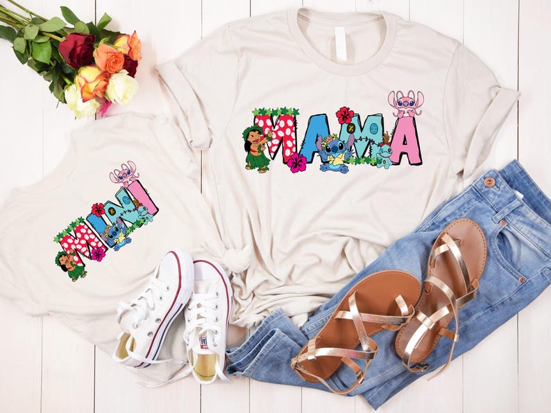 May include: Two white t-shirts with a cartoon character design. The larger shirt says "MAMA" and the smaller shirt says "MINI". Both shirts feature a cartoon character with blue and pink accents. The shirts are laid out on a white surface with a pair of jeans and sandals.