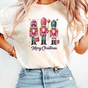 May include: A cream-coloured t-shirt with a festive design of three nutcracker soldiers in pink and teal uniforms. The words "Merry Christmas" are written in a cursive font below the soldiers, with gold star accents.