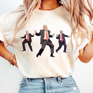 May include: Cream-coloured t-shirt with a graphic of three figures in black suits and red ties, in a dancing pose. The figures have blonde hair and are in various dance positions. The t-shirt is worn by a person in blue jeans.