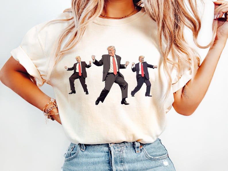 May include: Cream-coloured t-shirt with a graphic of three figures in black suits and red ties, in a dancing pose. The figures have blonde hair and are in various dance positions. The t-shirt is worn by a person in blue jeans.