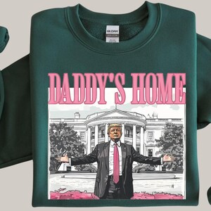 May include: Dark green sweatshirt with the text "DADDY'S HOME" in pink letters above an illustration of a man in a suit with the White House in the background. The man has his arms outstretched.