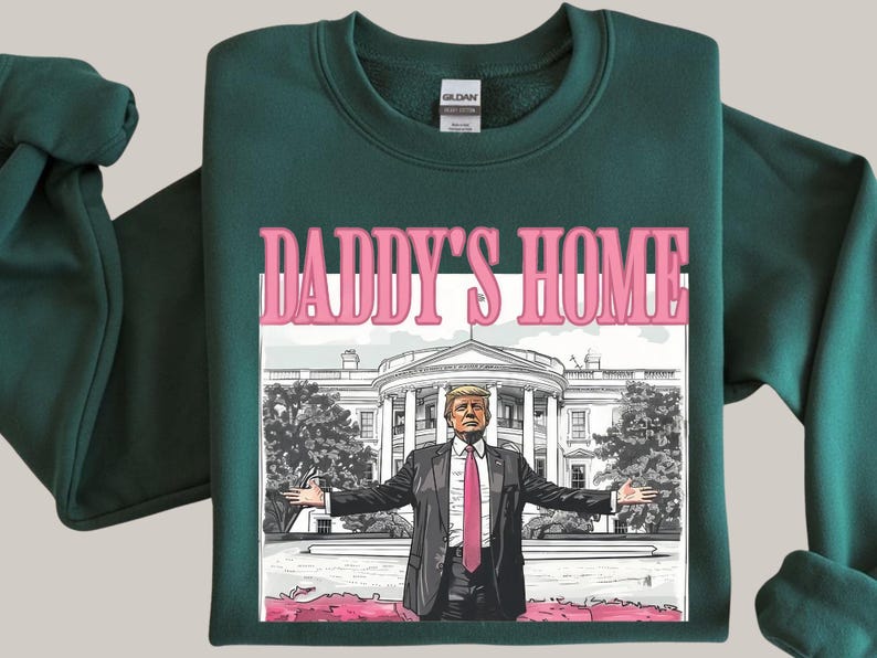 May include: Dark green sweatshirt with the text "DADDY'S HOME" in pink letters above an illustration of a man in a suit with the White House in the background. The man has his arms outstretched.