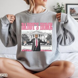 May include: Athletic heather gray sweatshirt with a pink graphic that says "DADDY'S HOME". The graphic features a cartoon illustration of a man in a suit with the White House in the background.