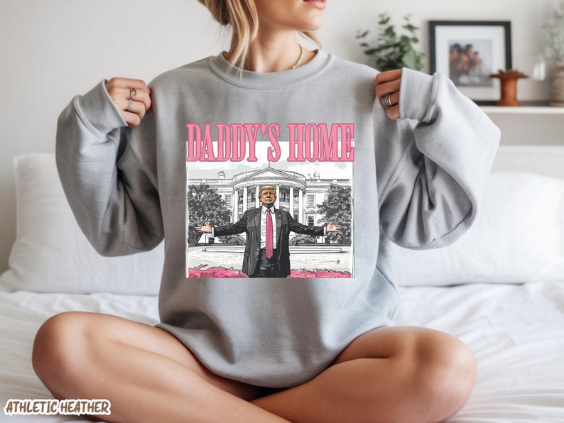 May include: Athletic heather gray sweatshirt with a pink graphic that says "DADDY'S HOME". The graphic features a cartoon illustration of a man in a suit with the White House in the background.