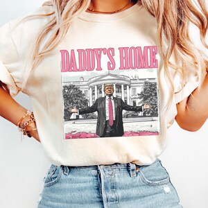 May include: Cream-colored t-shirt with the text "DADDY'S HOME" in pink. The graphic features a cartoon image of a man in a suit with open arms in front of the White House. The shirt is worn by a person wearing blue jeans.