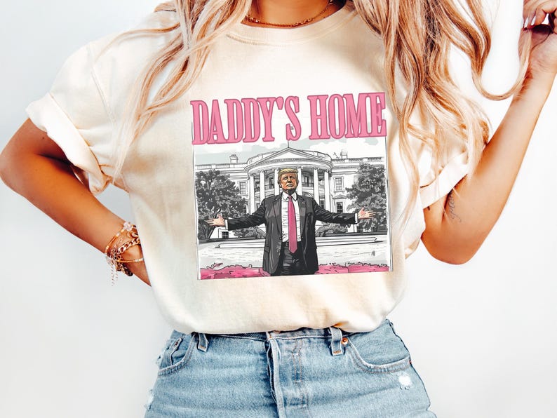 May include: Cream-colored t-shirt with the text "DADDY'S HOME" in pink. The graphic features a cartoon image of a man in a suit with open arms in front of the White House. The shirt is worn by a person wearing blue jeans.