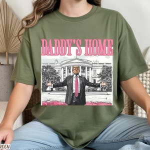 May include: Military green t-shirt with a graphic that says "DADDY'S HOME" in pink letters. The graphic features a man in a suit with his arms outstretched in front of the White House.
