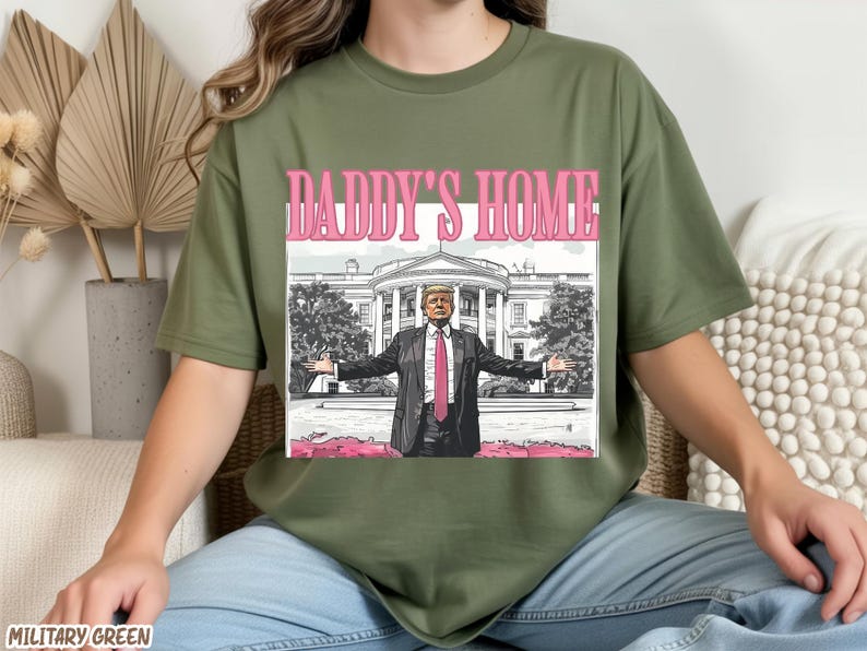 May include: Military green t-shirt with a graphic that says "DADDY'S HOME" in pink letters. The graphic features a man in a suit with his arms outstretched in front of the White House.