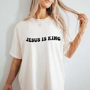 Puede incluir: Una camiseta blanca con el texto "JESUS IS KING" impreso en negro.