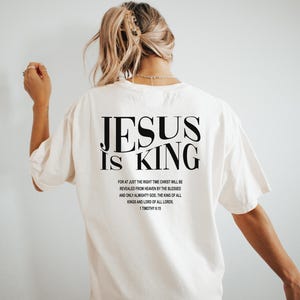 Puede incluir: Camiseta blanca con texto negro que dice "JESUS IS KING" y un vers&iacute;culo b&iacute;blico de 1 Timoteo 6:15.
