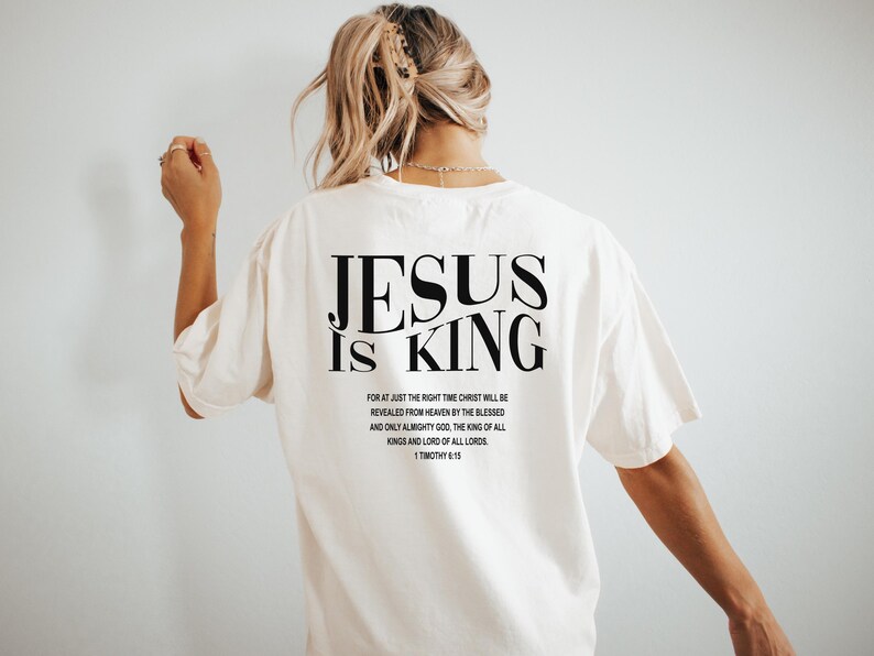 Puede incluir: Camiseta blanca con texto negro que dice "JESUS IS KING" y un vers&iacute;culo b&iacute;blico de 1 Timoteo 6:15.