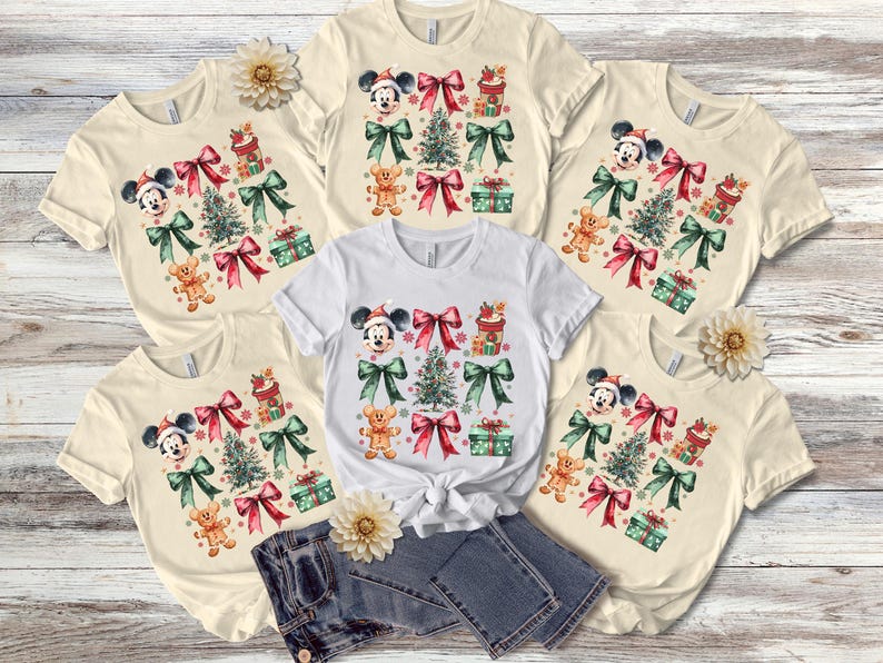 May include: Five cream-colored t-shirts with a Christmas theme featuring Mickey Mouse, a Christmas tree, presents, and bows. The shirts are arranged in a circle with a pair of blue jeans in the center.