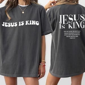 Puede incluir: Una camiseta gris oscura de gran tama&ntilde;o con el texto "JESUS IS KING" en blanco en la parte delantera y un texto m&aacute;s largo en blanco en la parte posterior. El texto en la parte posterior dice: "FOR AT JUST THE RIGHT TIME CHRIST WILL BE REVEALED FROM HEAVEN BY THE BLESSED AND ONLY ALMIGHTY GOD, THE KING OF ALL KINGS AND LORD OF ALL LORDS. 1 TIMOTHY 6:15"