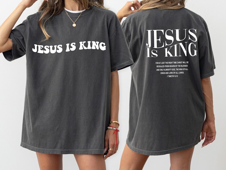 Puede incluir: Una camiseta gris oscura de gran tama&ntilde;o con el texto "JESUS IS KING" en blanco en la parte delantera y un texto m&aacute;s largo en blanco en la parte posterior. El texto en la parte posterior dice: "FOR AT JUST THE RIGHT TIME CHRIST WILL BE REVEALED FROM HEAVEN BY THE BLESSED AND ONLY ALMIGHTY GOD, THE KING OF ALL KINGS AND LORD OF ALL LORDS. 1 TIMOTHY 6:15"
