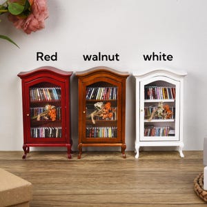 May include: Three miniature wooden bookcases in red, walnut, and white. Each bookcase has three shelves and glass doors. The shelves are filled with miniature books.