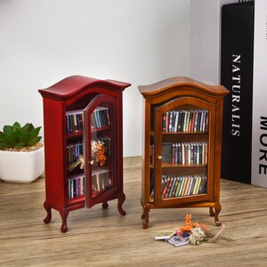 May include: Two miniature wooden bookcases, one red and one brown, with glass doors and shelves filled with miniature books. The bookcases have ornate details and are on a wooden surface.