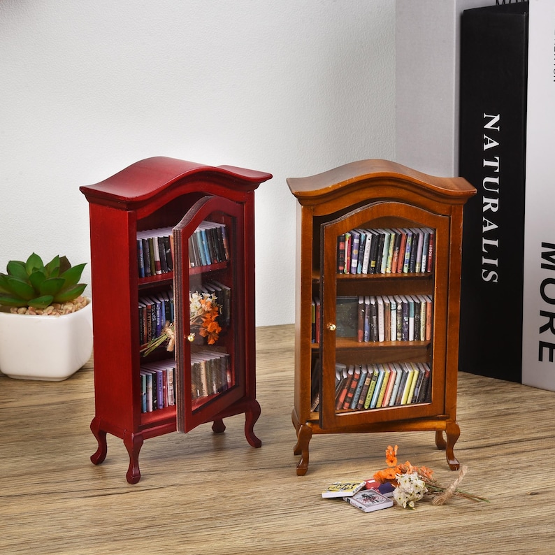 May include: Two miniature wooden bookcases, one red and one brown, with glass doors and shelves filled with miniature books. The bookcases have ornate details and are on a wooden surface.