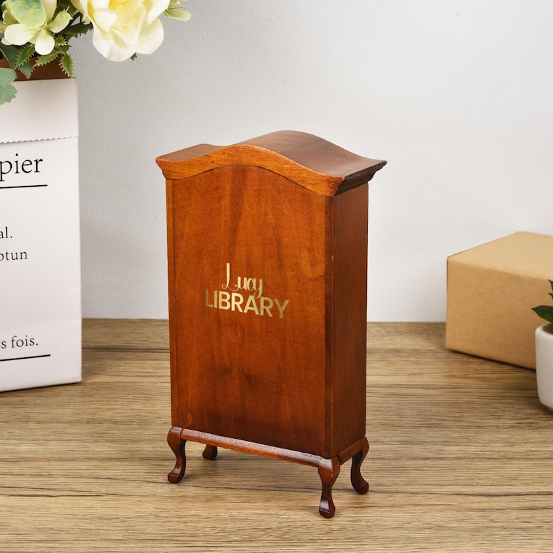May include: A wooden dollhouse-sized bookcase with a curved top and decorative feet. The bookcase is brown and has the words "Lucy Library" printed on the front in gold lettering.