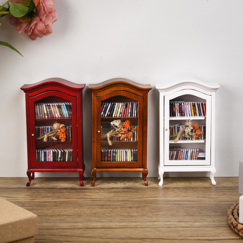 May include: Three miniature wooden bookcases with glass doors. The bookcases are painted red, brown, and white. Each bookcase has three shelves filled with miniature books. A small bouquet of dried flowers sits on the middle shelf of each bookcase.