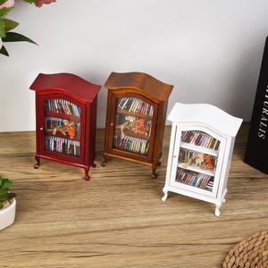 May include: Three miniature wooden bookcases, one red, one brown, and one white, each with glass doors and shelves filled with miniature books. The bookcases are on a wooden surface.