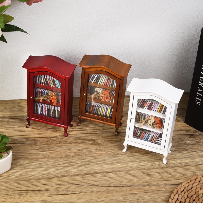 May include: Three miniature wooden bookcases, one red, one brown, and one white, each with glass doors and shelves filled with miniature books. The bookcases are on a wooden surface.