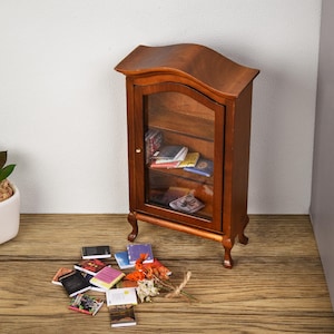 May include: A miniature wooden bookcase with glass doors and shelves filled with miniature books. The bookcase has a curved top and sits on four legs. There are several miniature books scattered on the floor in front of the bookcase.