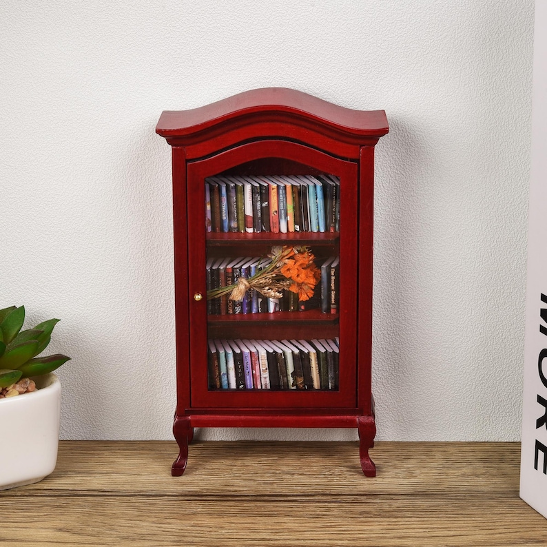 May include: A miniature red wooden bookcase with glass doors and shelves filled with books. A bouquet of orange flowers is placed on the top shelf.