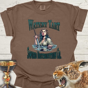Puede incluir: Una camiseta marrón con un diseño gráfico de una mujer sosteniendo una espada en un cuerpo de agua. El texto "Watery Tart Sword Distribution Co." está impreso en la camiseta.
