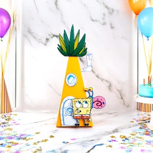 May include: A yellow pineapple-shaped decoration featuring a cartoon of SpongeBob SquarePants and his snail, Gary. The decoration has a green and yellow top, a porthole window, and a door. Balloons and a cake are in the background.