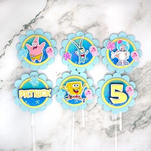 May include: Set of six cupcake toppers featuring characters from the animated series SpongeBob SquarePants. Each topper has a blue scalloped edge, a yellow circle, and a character image. Characters include Patrick, Plankton, Sandy, SpongeBob, and a number 5.