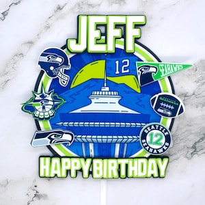 Könnte beinhalten: Ein grüner, weißer und blauer Tortendekor mit dem Namen "JEFF" und den Worten "Happy Birthday". Der Topper ist im Seattle Seahawks-Stil mit einem Space Needle, einem Fußball und einer Flagge.